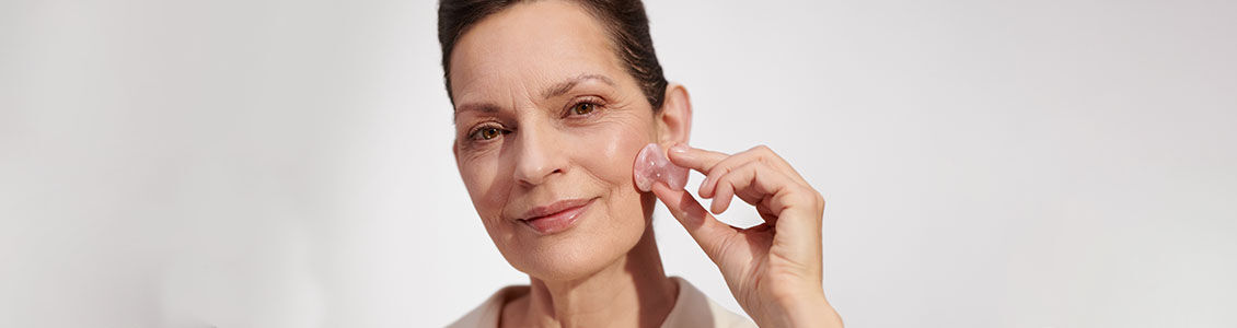 The image shows the face of an older woman gently massaging her face with the L'OCCITANE Massage tool