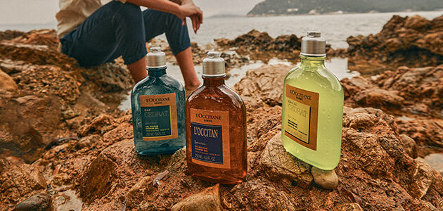 Man sitting on beach with L'OCCITANE Cap Cedrat Shower Gel, L'OCCITANE Eau de Cedrat Shower Gel & L'Occitan Shower Gel
