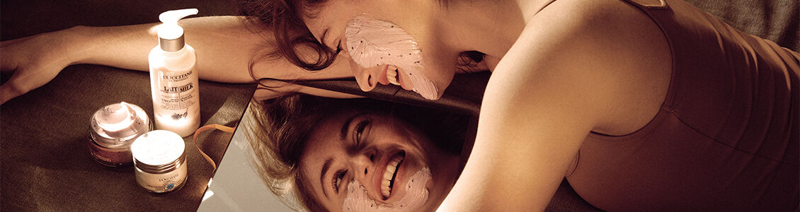 Woman looking in mirror in order to apply L'OCCITANE Soothing Face Mask to face.